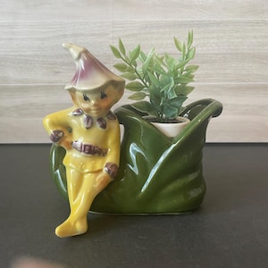 May include: A ceramic planter featuring a yellow elf figure with a purple-tipped hat sitting on the edge of a dark green, leaf-shaped vessel. The elf has painted facial features and a belt. A small green artificial plant is inside the planter.