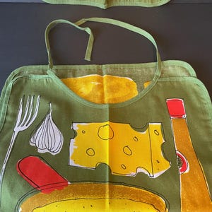 May include: An olive green apron featuring a graphic print of a white fork, a garlic bulb, a yellow slice of Swiss cheese, a red spatula, a yellow bowl of food, and a yellow bottle with a red cap. The apron has a neck strap and hemmed edges.