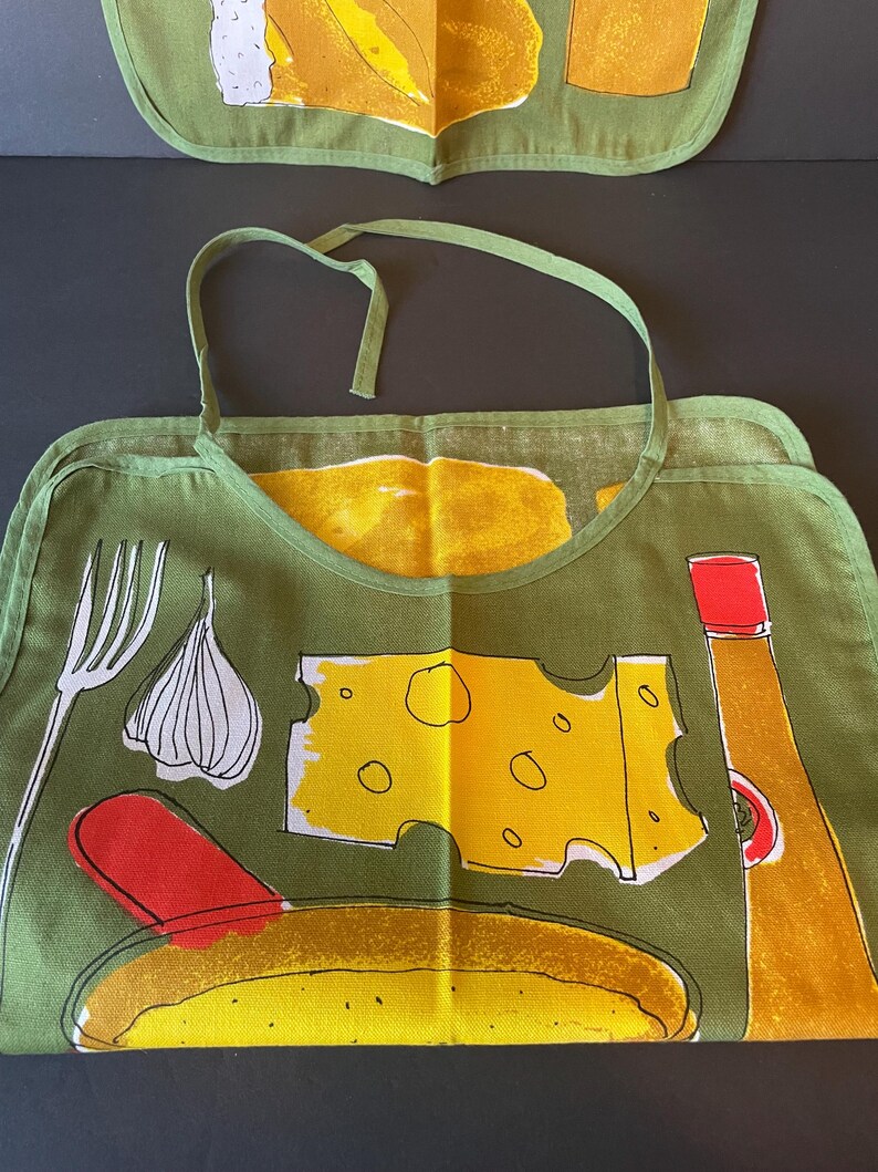 May include: An olive green apron featuring a graphic print of a white fork, a garlic bulb, a yellow slice of Swiss cheese, a red spatula, a yellow bowl of food, and a yellow bottle with a red cap. The apron has a neck strap and hemmed edges.