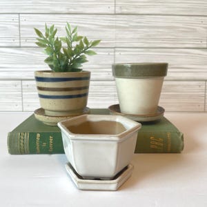 May include: Three ceramic planters with matching saucers. One is striped with blue and brown bands, another is cream-colored with a green rim, and the third is a white, geometric shape. A small green plant sits in the striped planter.