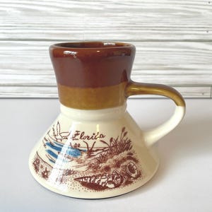 May include: A ceramic coffee mug with a brown top, tan middle, and cream-colored base. The base features a brown illustration of a Florida landscape with the word "Florida" in script. The mug has a curved handle.