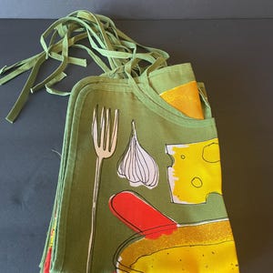May include: A folded olive green apron featuring a graphic print of a white fork, a white garlic bulb, a yellow block of Swiss cheese, and a red and yellow food item. The apron includes long green fabric ties at the top for securing around the waist.