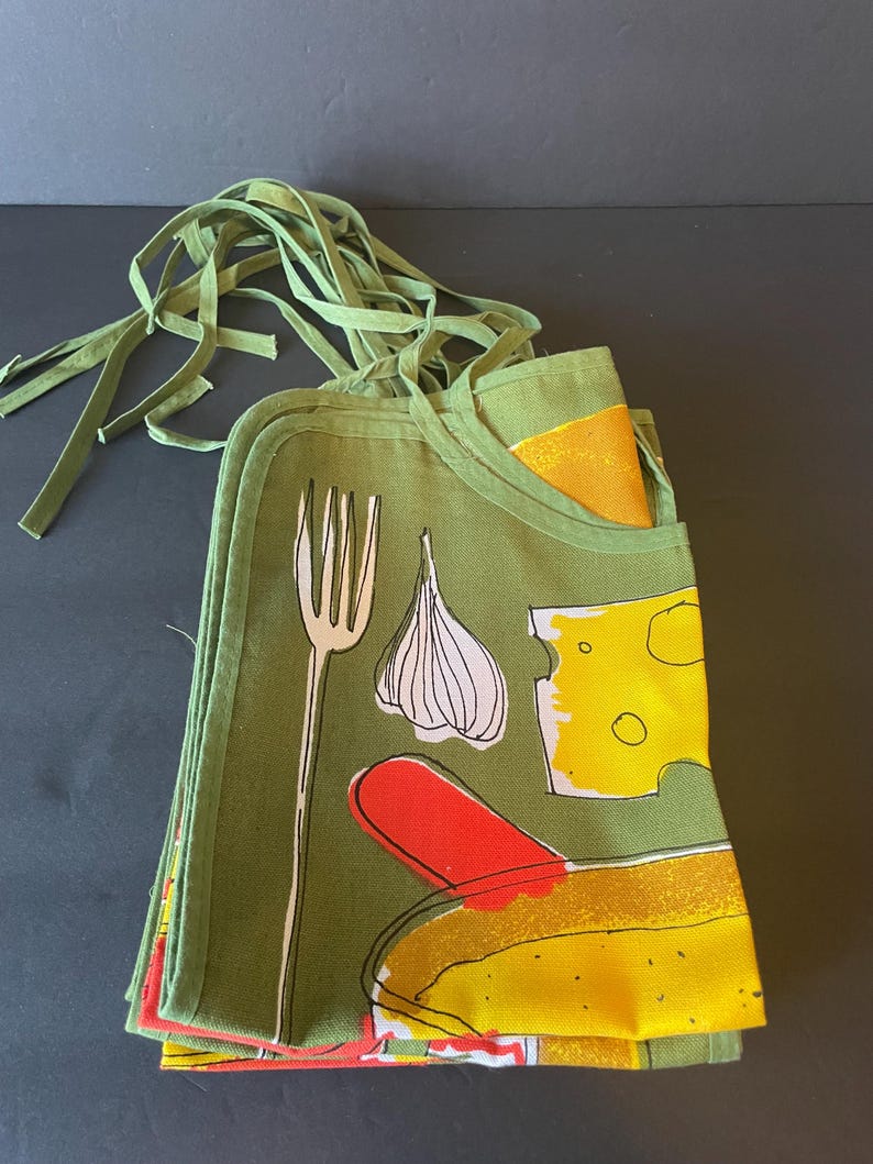 May include: A folded olive green apron featuring a graphic print of a white fork, a white garlic bulb, a yellow block of Swiss cheese, and a red and yellow food item. The apron includes long green fabric ties at the top for securing around the waist.