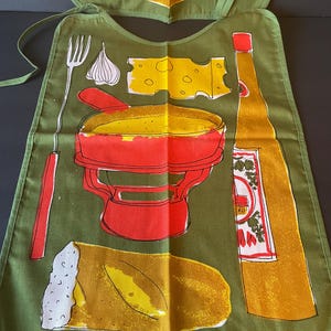 May include: An olive green apron featuring a graphic print of a red fondue pot, a long fork, a wedge of yellow Swiss cheese, a clove of garlic, a loaf of bread, and a tall yellow bottle with a red cap and a label with red and green illustrations.