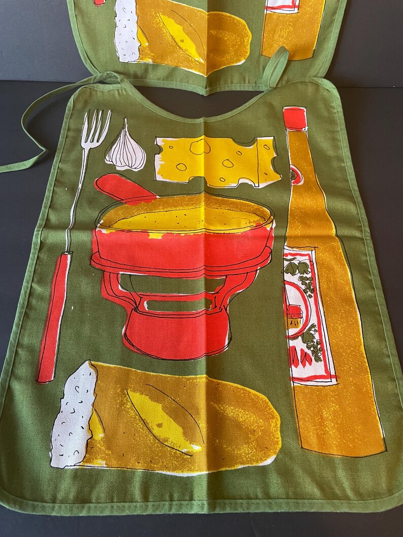 May include: An olive green apron featuring a graphic print of a red fondue pot, a long fork, a wedge of yellow Swiss cheese, a clove of garlic, a loaf of bread, and a tall yellow bottle with a red cap and a label with red and green illustrations.