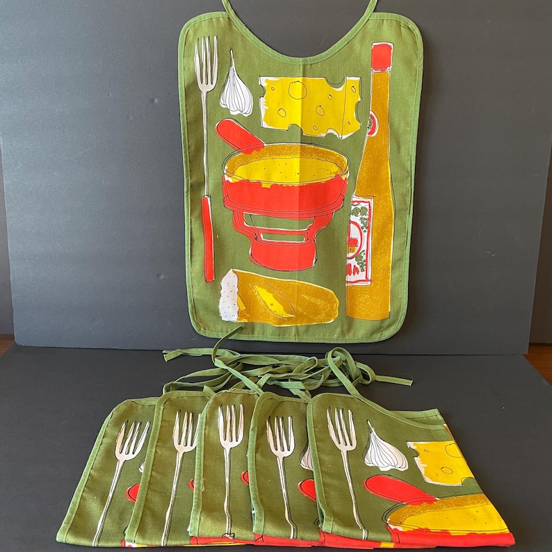 May include: A set of olive green fabric bibs featuring a screen-printed graphic of a fondue pot, a block of Swiss cheese, a bottle of hot sauce, a clove of garlic, a piece of bread, and a long-handled fork, all tied with matching green fabric strings.