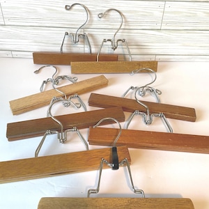 May include: A collection of vintage wooden skirt hangers with metal hooks and clips. The wooden bars vary in shades of brown, from light to dark. The metal components are silver-toned. The hangers are arranged on a white surface.