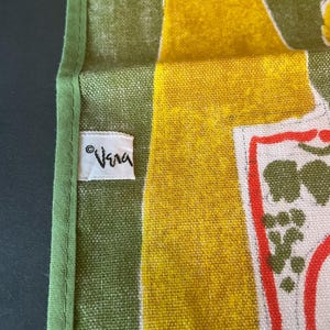 May include: A green and yellow textile featuring a white fabric tag with the text Vera printed in black. The fabric has a visible woven texture with a green border and a graphic pattern that includes red outlines and green shapes on a white background.
