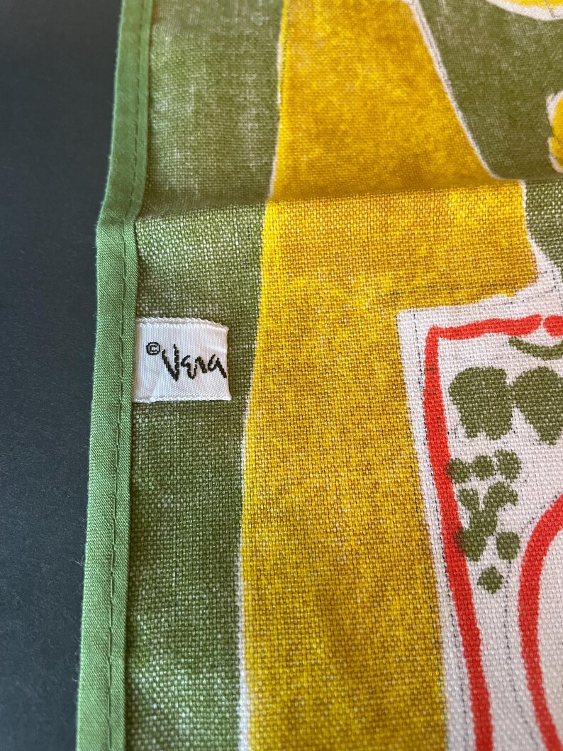 May include: A green and yellow textile featuring a white fabric tag with the text Vera printed in black. The fabric has a visible woven texture with a green border and a graphic pattern that includes red outlines and green shapes on a white background.