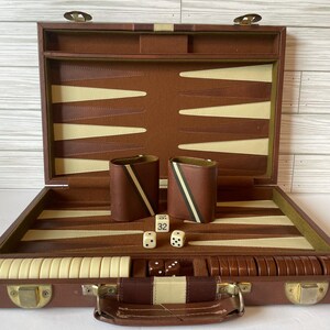 May include: Open brown leather backgammon set with ivory and brown playing surface. Includes dice, dice cups, and game pieces. The set is displayed open, revealing the interior game board and accessories.