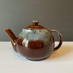 May include: A brown teapot with a white glaze. The teapot has a round body and a small spout. The handle is on the side of the teapot.