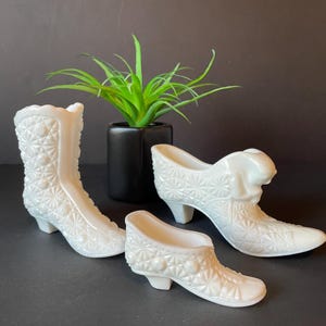 May include: Three white, decorative ceramic shoes and a boot with a textured, quilted pattern. The shoes are in different styles, including a high-heeled pump and a small slipper. A small, green plant in a black pot is in the background.
