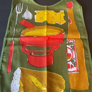 May include: An olive green apron featuring a graphic print of a red fondue pot, a yellow block of Swiss cheese, a long fork, a clove of garlic, a loaf of bread, and a tall yellow bottle with a red cap and a label depicting grapes and a house.