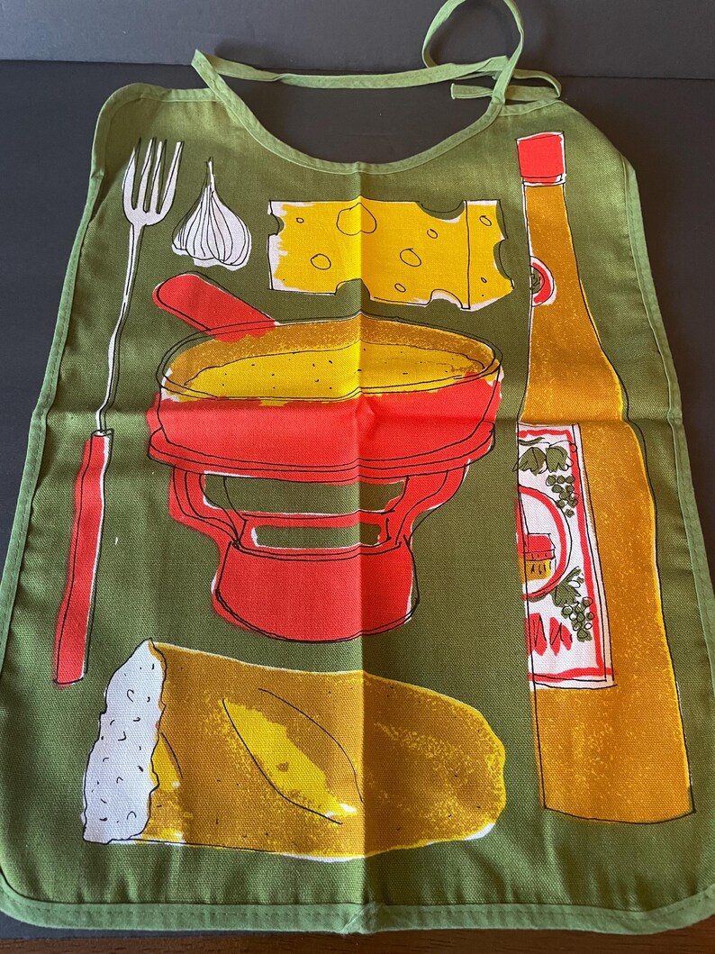 May include: An olive green apron featuring a graphic print of a red fondue pot, a yellow block of Swiss cheese, a long fork, a clove of garlic, a loaf of bread, and a tall yellow bottle with a red cap and a label depicting grapes and a house.
