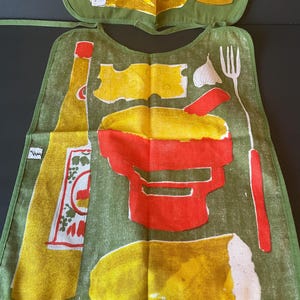 May include: A green fabric bib featuring a screen-printed design of a yellow bottle, a red mortar and pestle, a piece of yellow cheese, a white garlic clove, a white fork, and a loaf of bread. A small white tag with black text is on the left side.