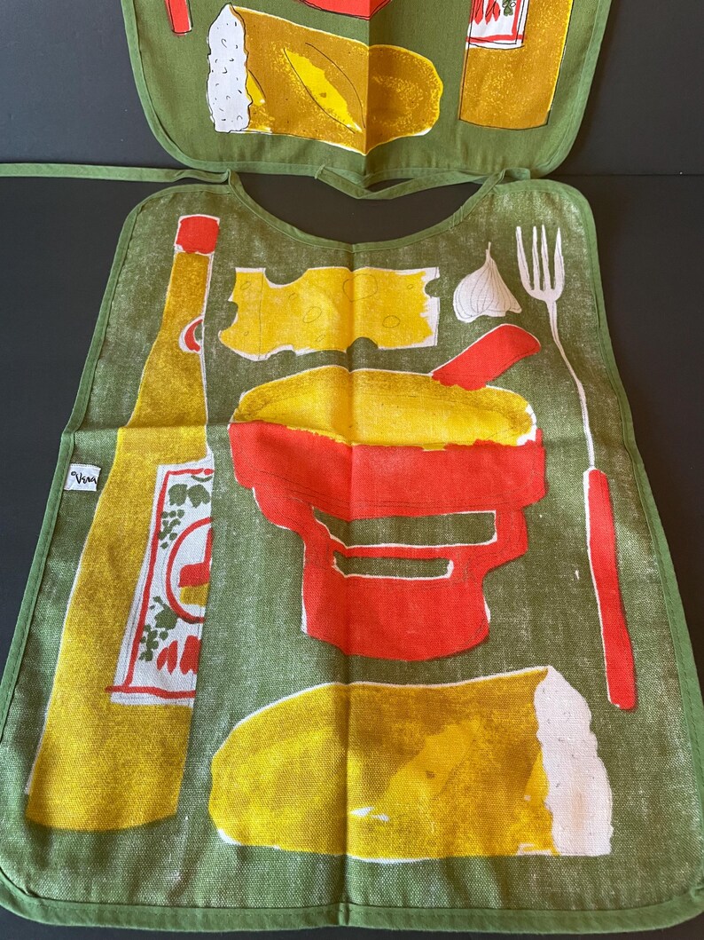 May include: A green fabric bib featuring a screen-printed design of a yellow bottle, a red mortar and pestle, a piece of yellow cheese, a white garlic clove, a white fork, and a loaf of bread. A small white tag with black text is on the left side.