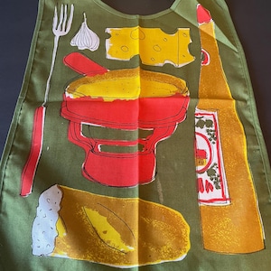May include: An olive green bib featuring a graphic print of a red fondue pot, a yellow piece of Swiss cheese, a loaf of bread, a garlic bulb, a fork, and a tall yellow bottle. The bib has a rounded neckline with fabric ties at the top corners.