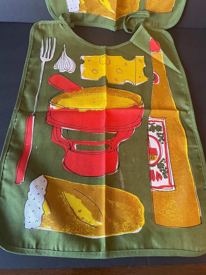 May include: An olive green bib featuring a graphic print of a red fondue pot, a yellow piece of Swiss cheese, a loaf of bread, a garlic bulb, a fork, and a tall yellow bottle. The bib has a rounded neckline with fabric ties at the top corners.
