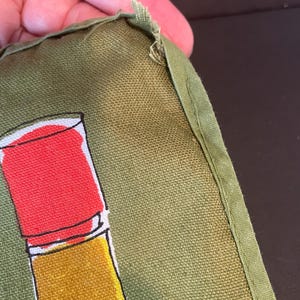 May include: An olive green fabric item featuring a graphic print of a red and yellow cylindrical object. The fabric has a visible woven texture and a stitched green border along the edge, showing slight fraying at the corner seam.