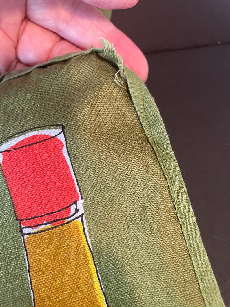 May include: An olive green fabric item featuring a graphic print of a red and yellow cylindrical object. The fabric has a visible woven texture and a stitched green border along the edge, showing slight fraying at the corner seam.