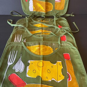 May include: A stack of olive green fabric bibs featuring a screen-printed culinary design. The pattern includes illustrations of a white fork, a garlic bulb, a yellow block of Swiss cheese, a red-capped bottle, and a loaf of bread in yellow and white.