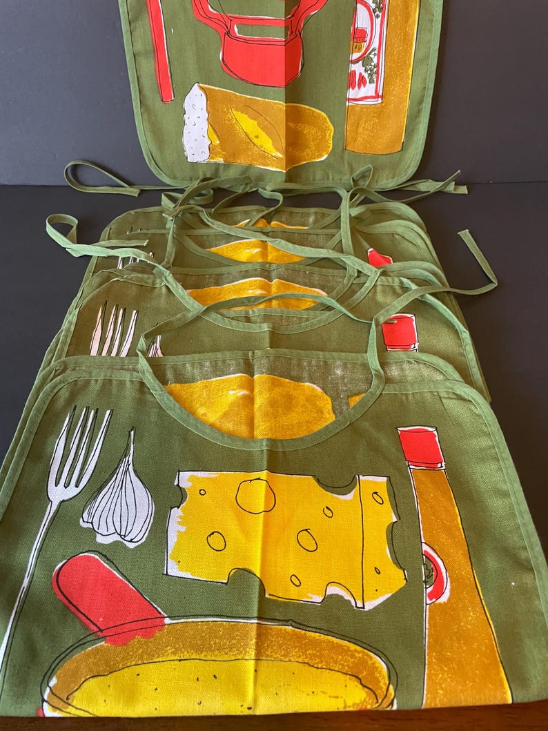 May include: A stack of olive green fabric bibs featuring a screen-printed culinary design. The pattern includes illustrations of a white fork, a garlic bulb, a yellow block of Swiss cheese, a red-capped bottle, and a loaf of bread in yellow and white.