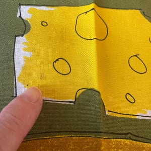 May include: A green fabric apron featuring a graphic print of a yellow block of Swiss cheese with black outlines and circular holes. The cheese design is set against a solid olive green background, with a textured weave visible on the fabric surface.