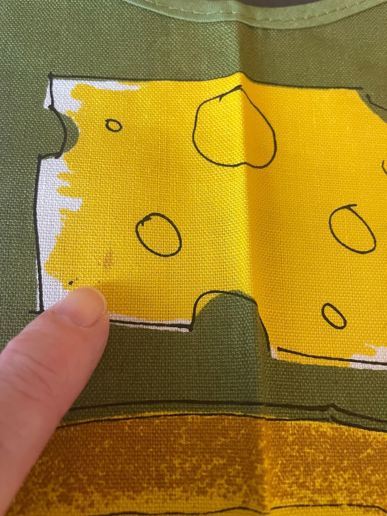 May include: A green fabric apron featuring a graphic print of a yellow block of Swiss cheese with black outlines and circular holes. The cheese design is set against a solid olive green background, with a textured weave visible on the fabric surface.