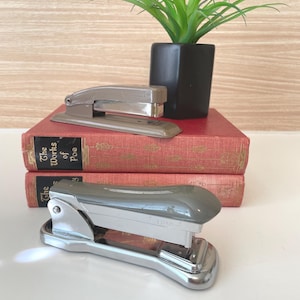 May include: Two vintage staplers, one gray and one silver, rest on a stack of red books with gold lettering. A small, black square pot holds a green plant, adding a touch of nature to the scene. The composition is set against a light wood-grain background.