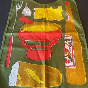 May include: An olive green apron featuring a graphic print of a red fondue pot, a yellow block of Swiss cheese, a clove of garlic, a long fondue fork, a loaf of crusty bread, and a tall yellow bottle with a white label containing red and green details.