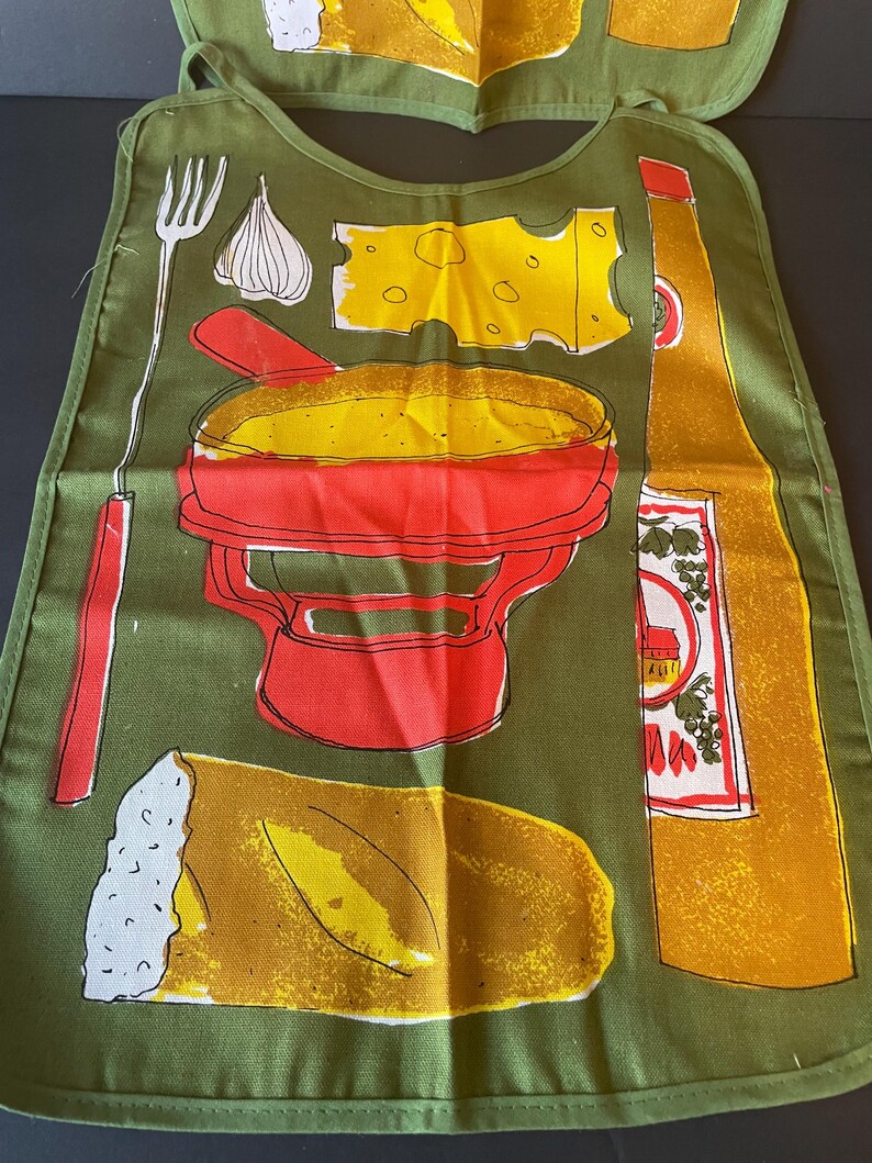 May include: An olive green apron featuring a graphic print of a red fondue pot, a yellow block of Swiss cheese, a clove of garlic, a long fondue fork, a loaf of crusty bread, and a tall yellow bottle with a white label containing red and green details.
