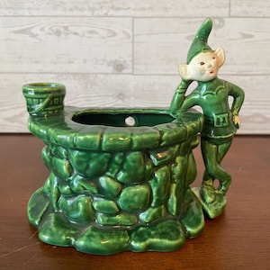 May include: A green ceramic planter shaped like a stone wishing well with a small bucket on the rim. A green-clad elf figure with a pale face and pointed hat leans against the side of the well. The structure has a glossy finish and a hollow center.