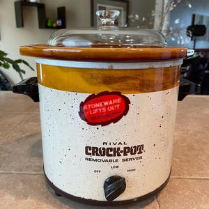 May include: A Rival Crock-Pot removable server with a cream-colored speckled body and a brown rim. It features a clear glass lid, black side handles, and a black control knob with settings for off, low, and high. A red sticker reads STONEWARE LIFTS OUT.