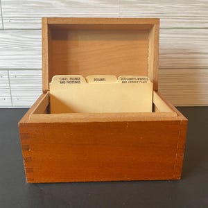 May include: A wooden recipe box with a hinged lid, containing recipe cards. The box is made of light brown wood and has dovetail joints. The recipe cards are organized with labeled dividers: Cakes, Fillings and Frostings, Desserts, and Doughnuts-Waffles and Griddle Cakes.