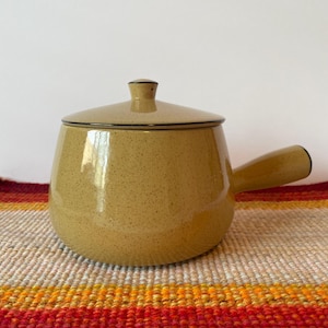 May include: A speckled, tan ceramic pot with a lid and a handle. The pot has a rounded shape and a dark rim. The lid has a small knob. The pot is sitting on a colorful, striped textile.