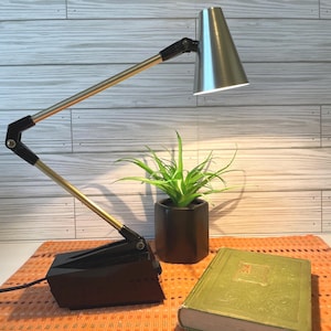 May include: A desk lamp with a black base, gold-toned adjustable arm, and a silver conical shade. The lamp is positioned next to a small green plant in a black hexagonal pot and a closed green hardcover book with gold embossed detailing.