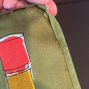 May include: An olive green fabric item featuring a graphic illustration of a lipstick tube with a bright red cap and a golden yellow base. The fabric has a visible woven texture and a stitched edge with a small frayed area at the top corner.