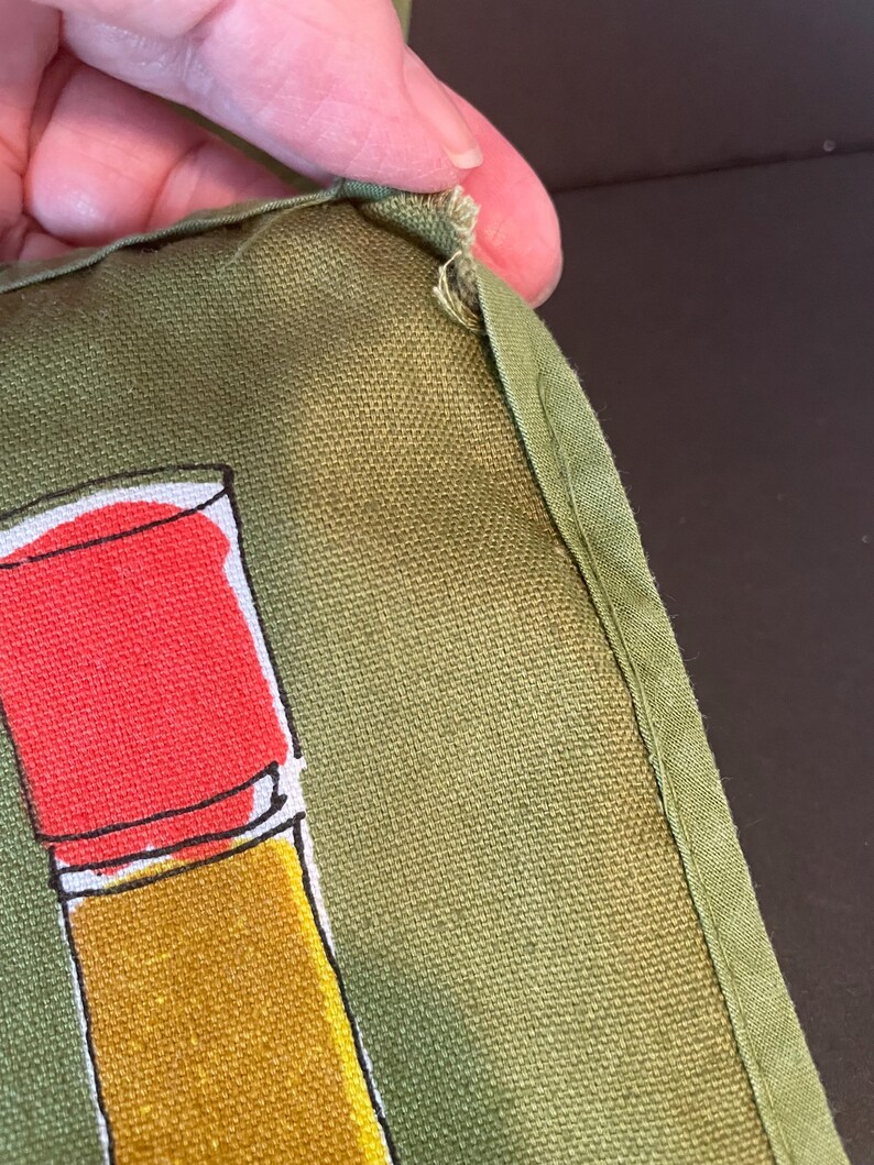 May include: An olive green fabric item featuring a graphic illustration of a lipstick tube with a bright red cap and a golden yellow base. The fabric has a visible woven texture and a stitched edge with a small frayed area at the top corner.