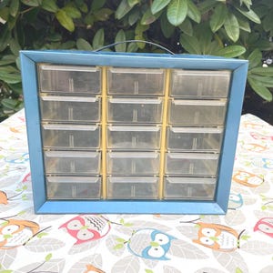 May include: A blue metal storage cabinet featuring fifteen clear plastic drawers arranged in a three-by-five grid. The unit includes a black handle on the top edge for portability and a yellow internal frame structure holding the drawers in place.