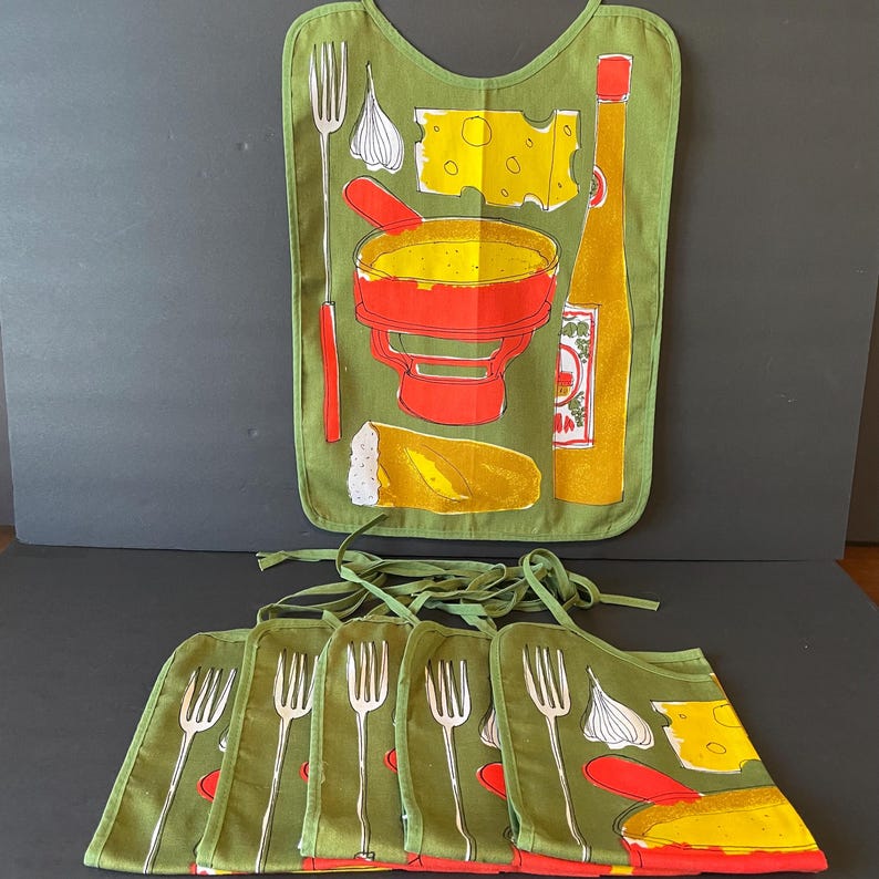 May include: A set of olive green bibs featuring a graphic print of a fondue pot, a piece of bread, a block of Swiss cheese, a garlic bulb, a fork, and a bottle of sauce. Each bib has fabric ties at the top for securing around the neck area.