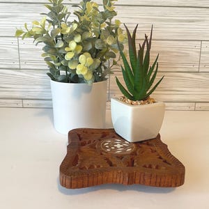 May include: A square wooden trivet with intricate carved patterns and a central circular inlay featuring a white floral design. A white pot containing a green leafy plant and a small white square planter with a green succulent sit on top.