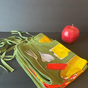 May include: A stack of folded olive green aprons featuring a graphic print of a white garlic bulb, a yellow block of cheese, and a red shape, accompanied by a single red apple. The aprons have long green fabric ties extending from the top edges.