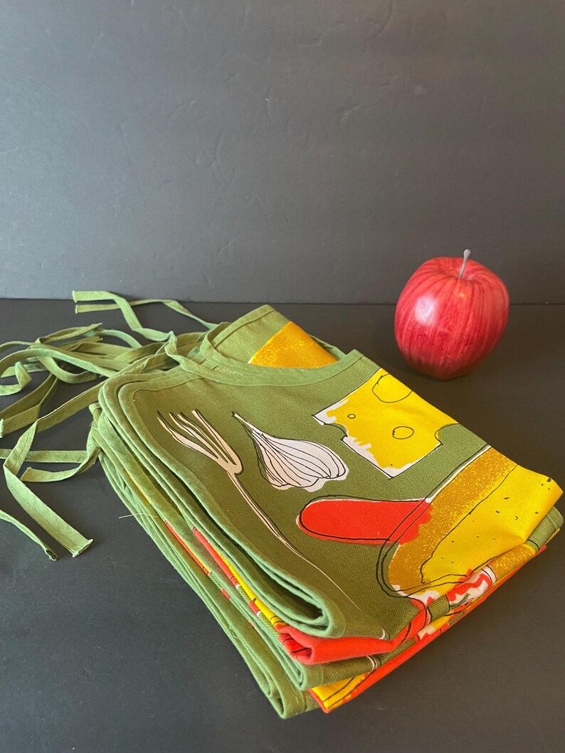 May include: A stack of folded olive green aprons featuring a graphic print of a white garlic bulb, a yellow block of cheese, and a red shape, accompanied by a single red apple. The aprons have long green fabric ties extending from the top edges.