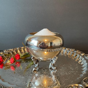 May include: A silver sugar bowl with a round body and three ornate feet, filled with white sugar, sitting on a silver tray with an ornate design.