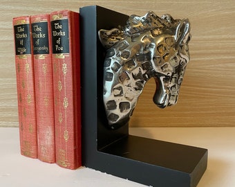 May include: A silver giraffe head bookend with a black base. The bookend is holding up three books with red covers. The books are titled "The Works of Wilde", "The Works of Dostoevsky", and "The Works of Poe".