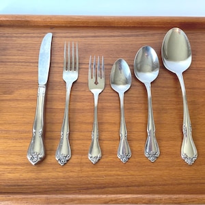 May include: A set of six silver-colored utensils, including a knife, two forks, and three spoons, arranged on a wooden tray. The utensils have decorative floral detailing on the handles. The tray is made of wood with a light brown finish.