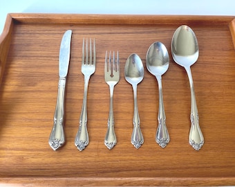 May include: A set of six silver-colored utensils, including a knife, two forks, and three spoons, arranged on a wooden tray. The utensils have decorative floral detailing on the handles. The tray is made of wood with a light brown finish.