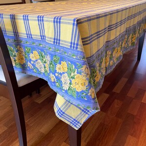 May include: A yellow and blue checkered tablecloth with a floral border. The tablecloth is draped over a wooden table with two chairs.