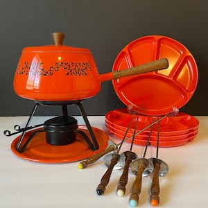 May include: A bright orange fondue pot with a black floral pattern, a wooden handle, and a matching lid. It sits on a black metal stand with a burner. The set includes four orange divided plates and four metal fondue forks with wooden handles.