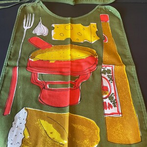 May include: An olive green adult bib featuring a graphic print of a red fondue pot, a piece of yellow cheese, a garlic bulb, a fork, a loaf of bread, and a tall bottle of oil. The design includes bold outlines and vibrant yellow and red accents.
